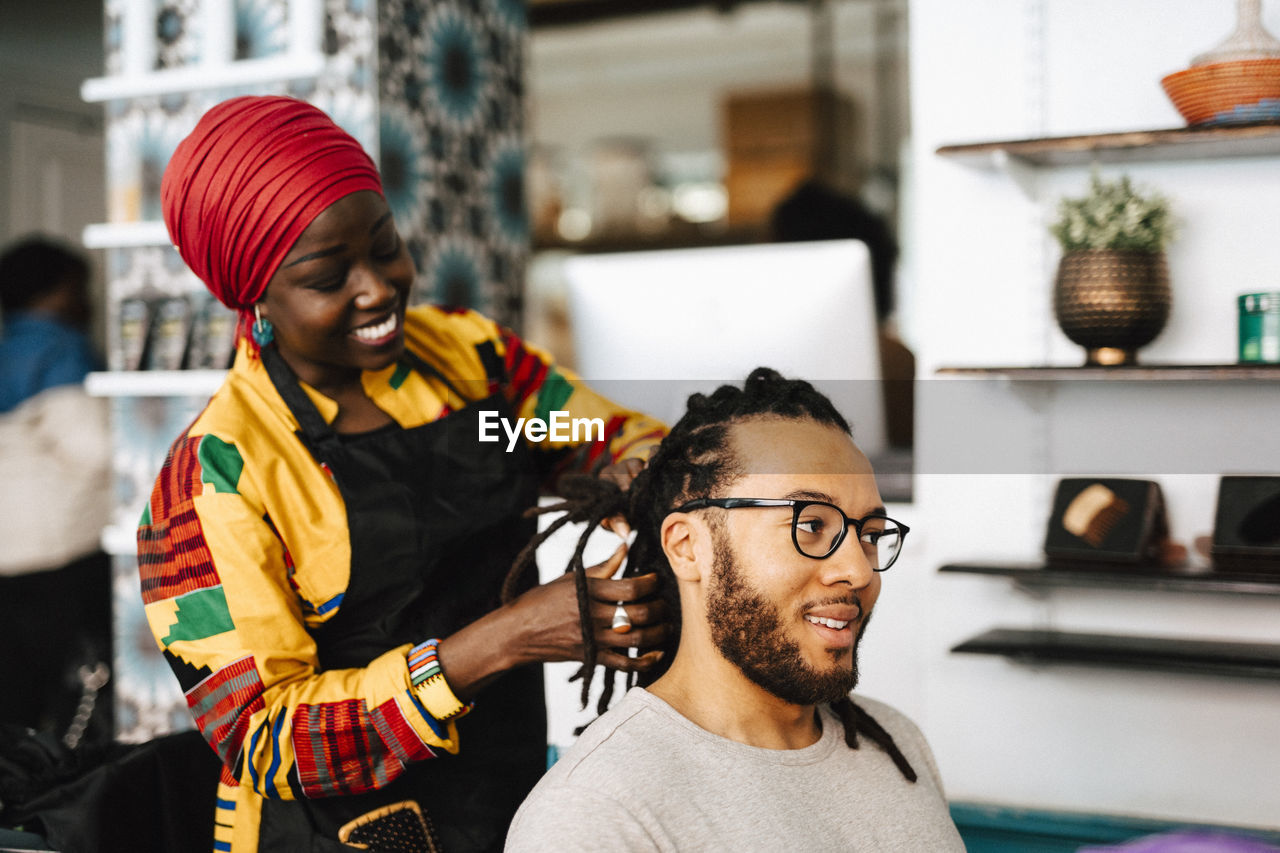 Smiling female barber making locs of male customer at salon