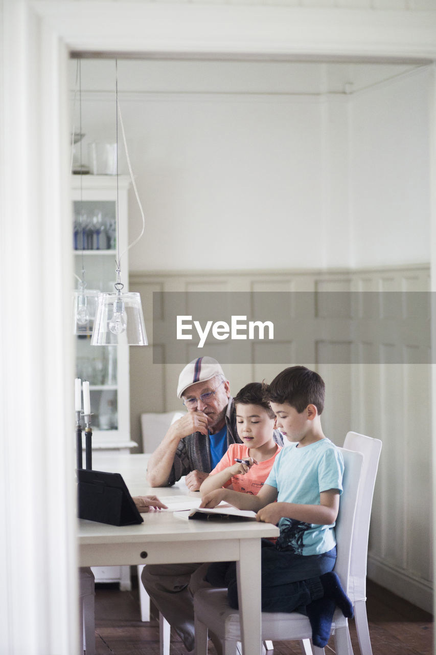 Grandfather sitting by twin grandsons reading book at table