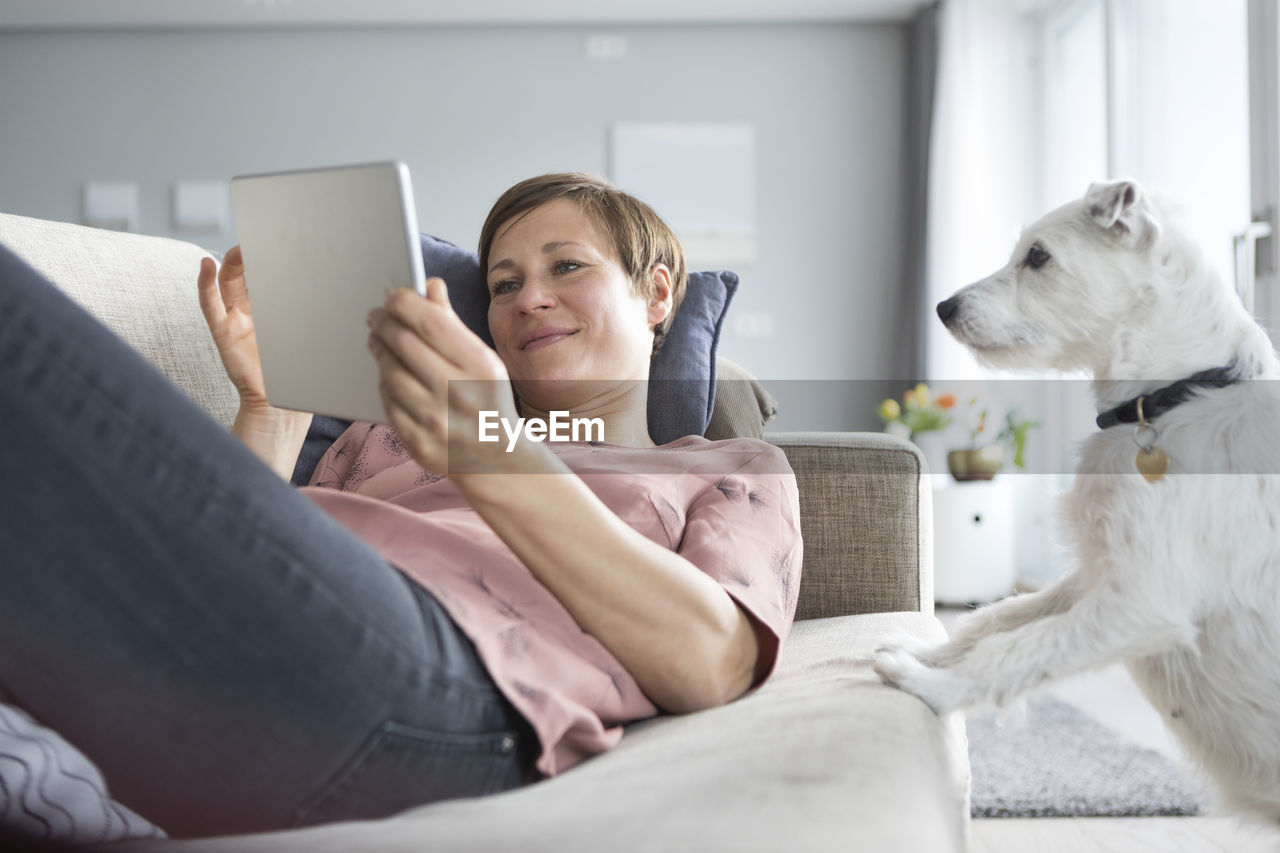 Portrait of smiling woman lying on the couch using tablet while dog watching her