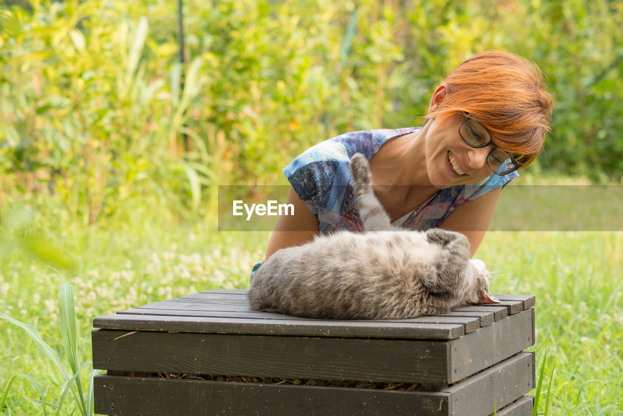 Smiling woman playing with cat on wooden seat at yard