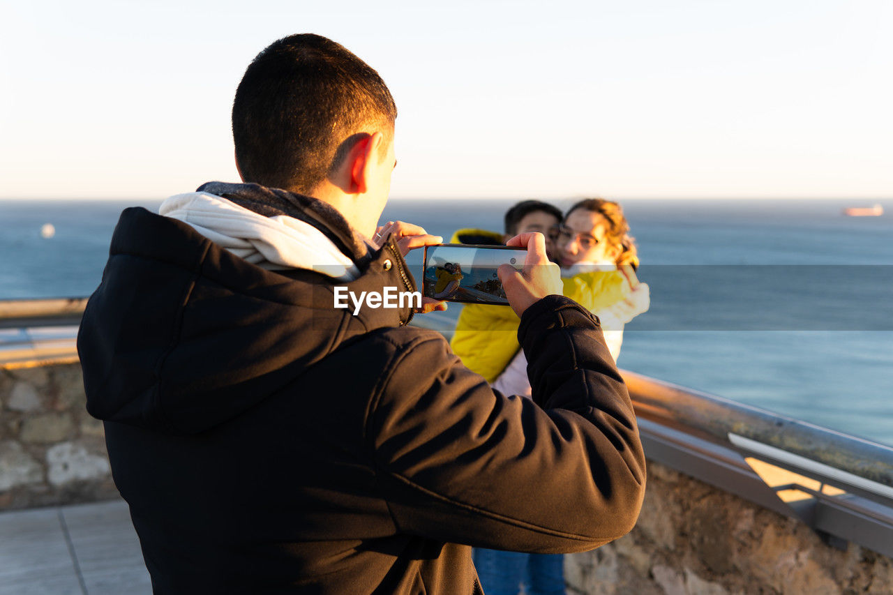A man takes a picture of his wife and son with his cell phone. the sea is in the background.