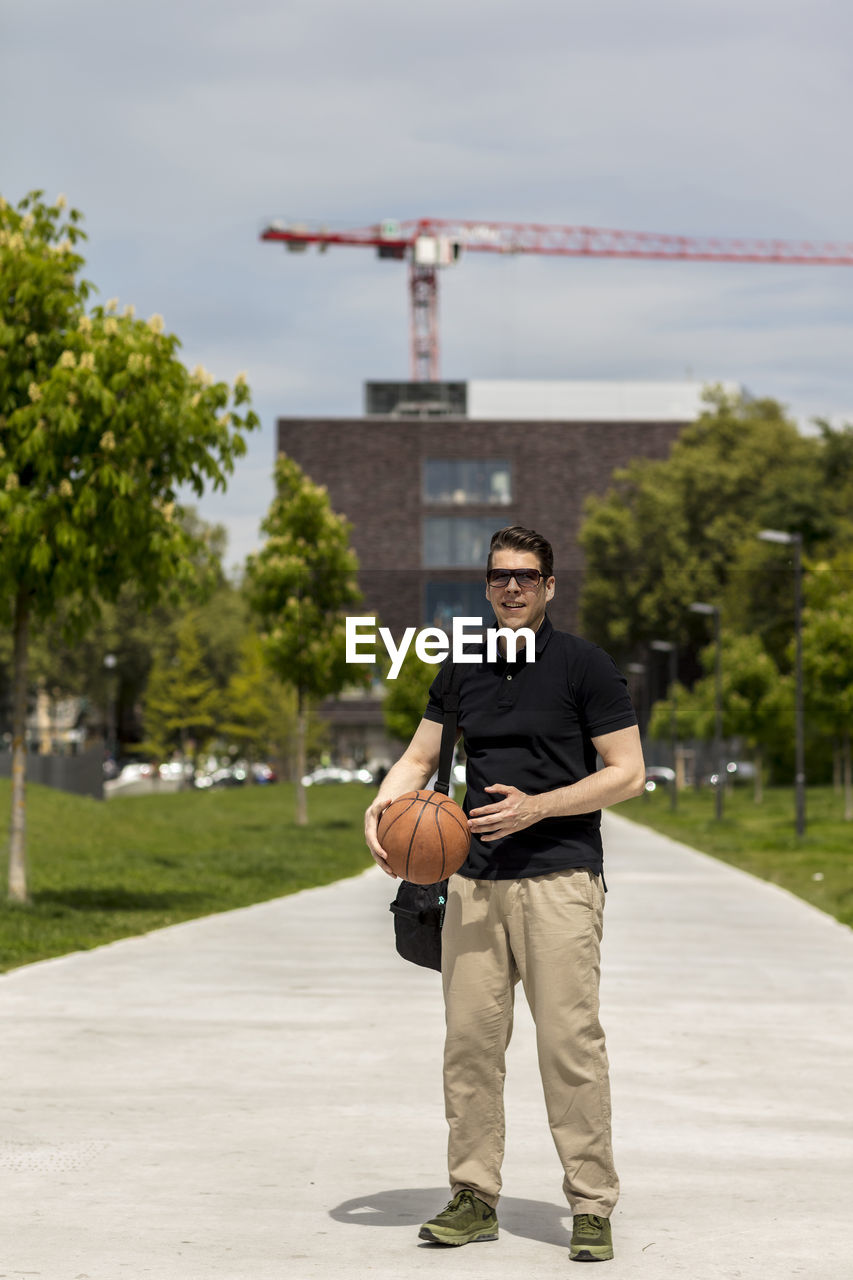 Full length of man holding basketball while standing on footpath against sky