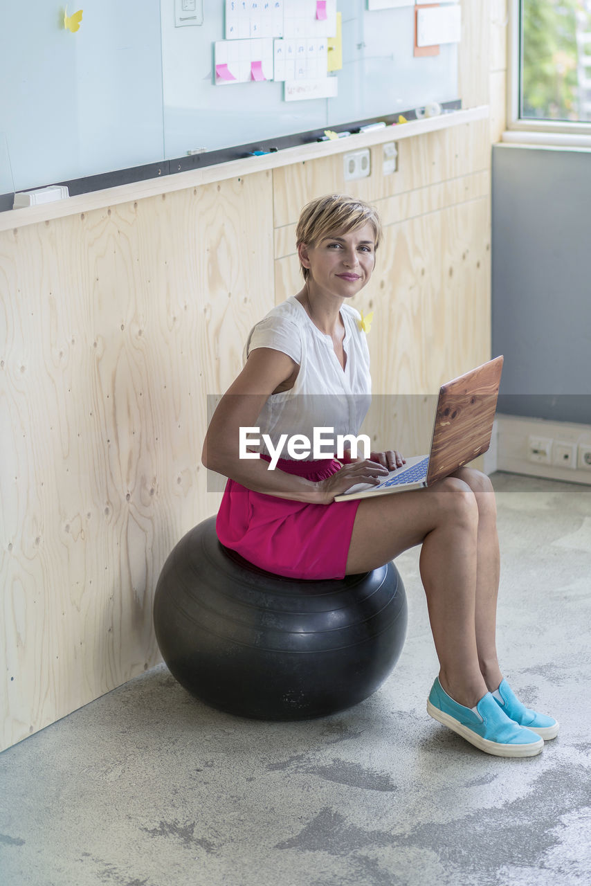 Smiling businesswoman sitting on fitness ball using laptop