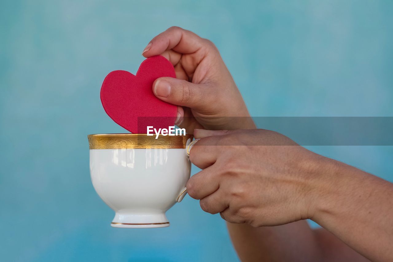 Close-up of hand holding heart shape in tea cup against blue wall