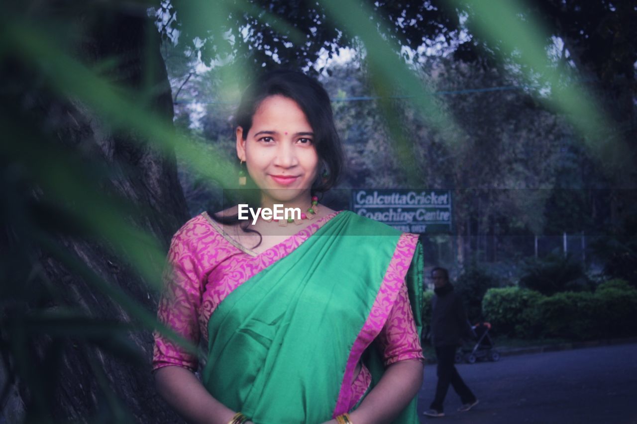 PORTRAIT OF SMILING YOUNG WOMAN STANDING BY TREES