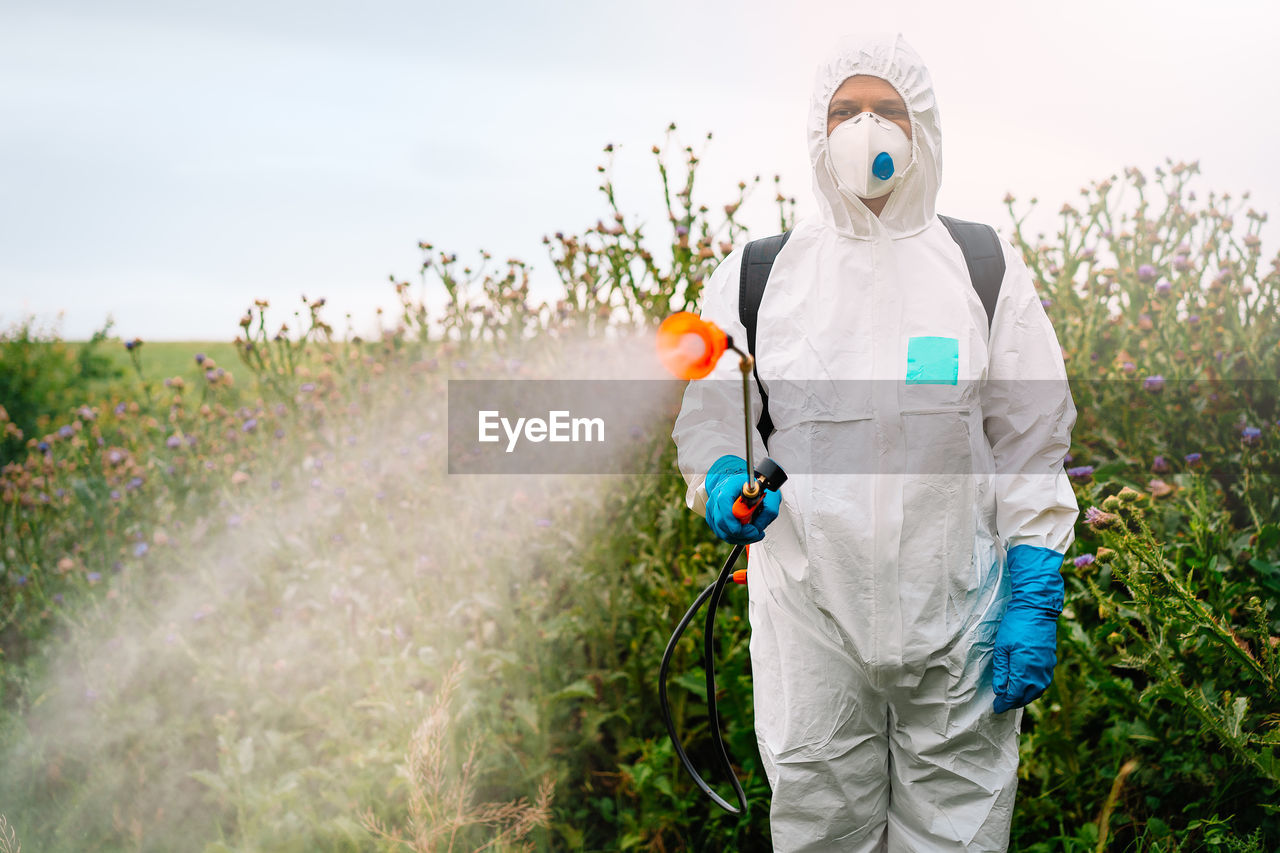 Person in protective workwear and mask spraying herbicide on thistle field