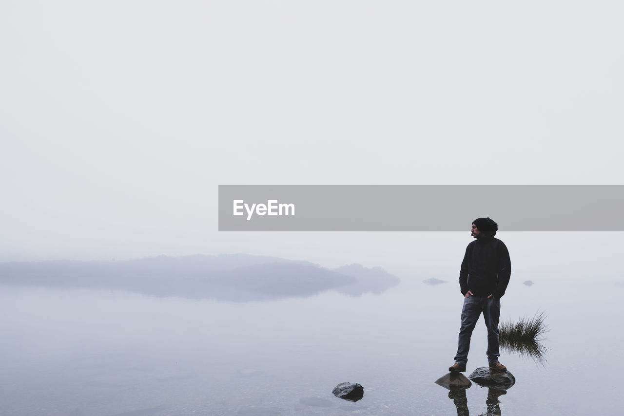 Man standing on rocks in lake against clear sky during foggy weather