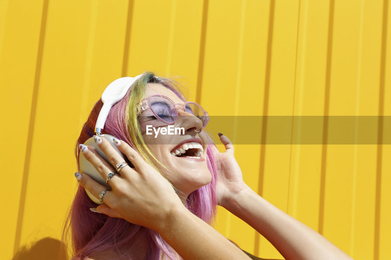 Happy woman wearing sunglasses enjoying music listening through wireless headphones in front of yellow wall