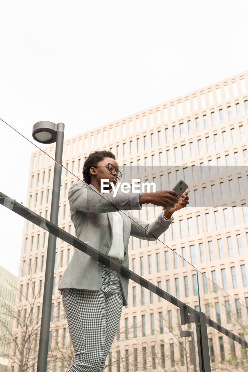 From below of young african american female in eyeglasses and classy outfit browsing on smartphone standing near office building leaning on glass barrier