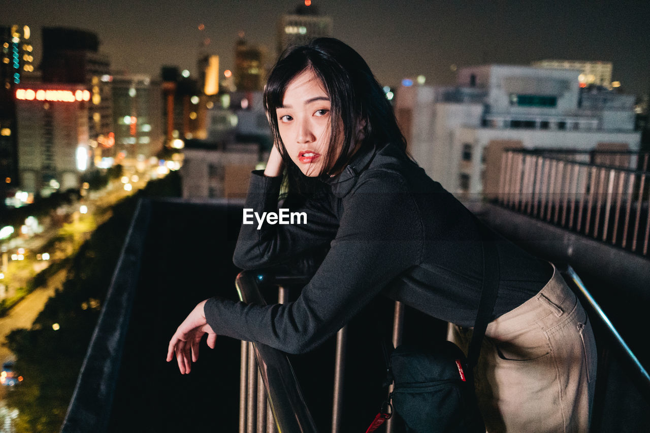 PORTRAIT OF YOUNG WOMAN STANDING ON ILLUMINATED CITY AT NIGHT