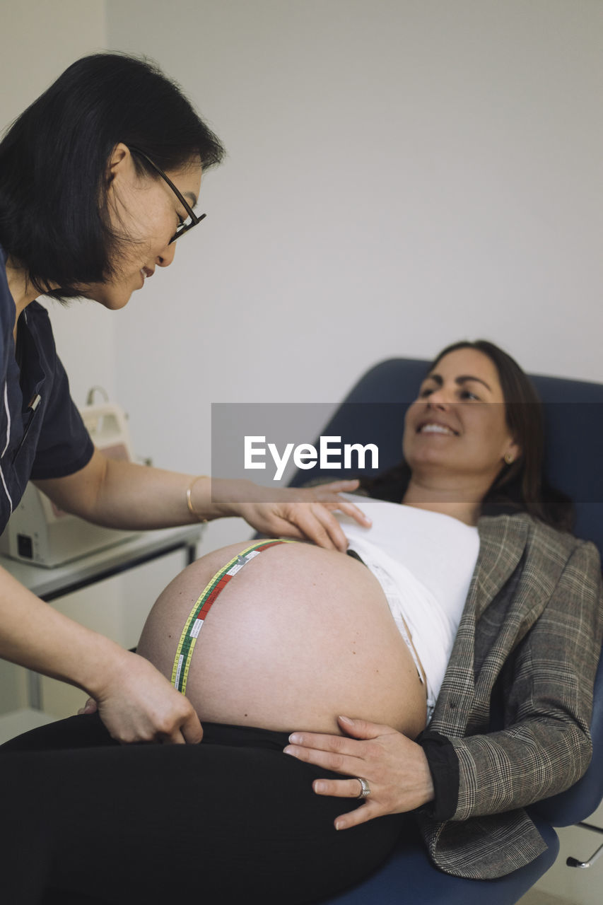 Smiling doctor measuring abdomen of pregnant patient lying on examination table in medical clinic