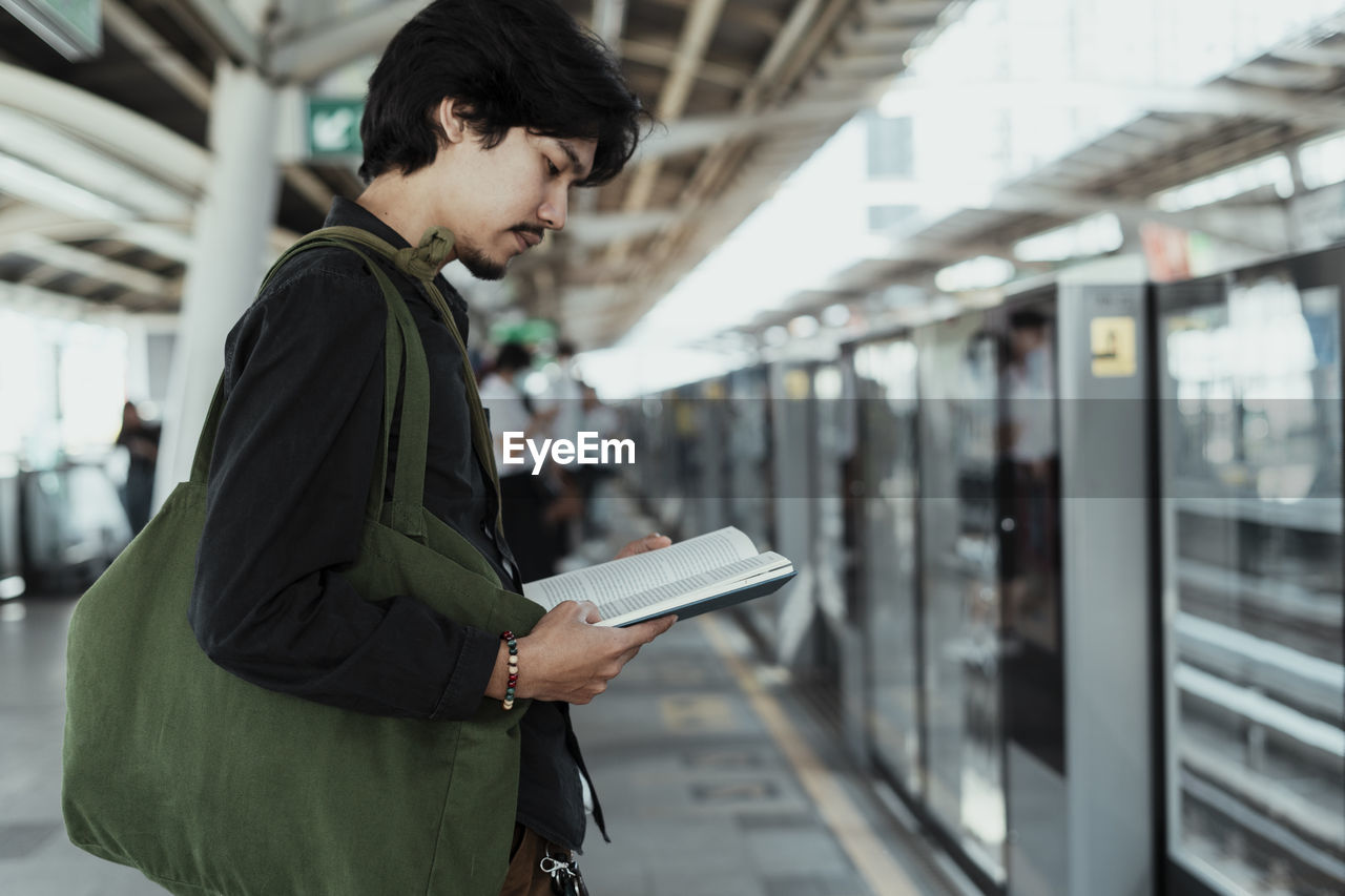 Side view of man reading book while standing at railroad station