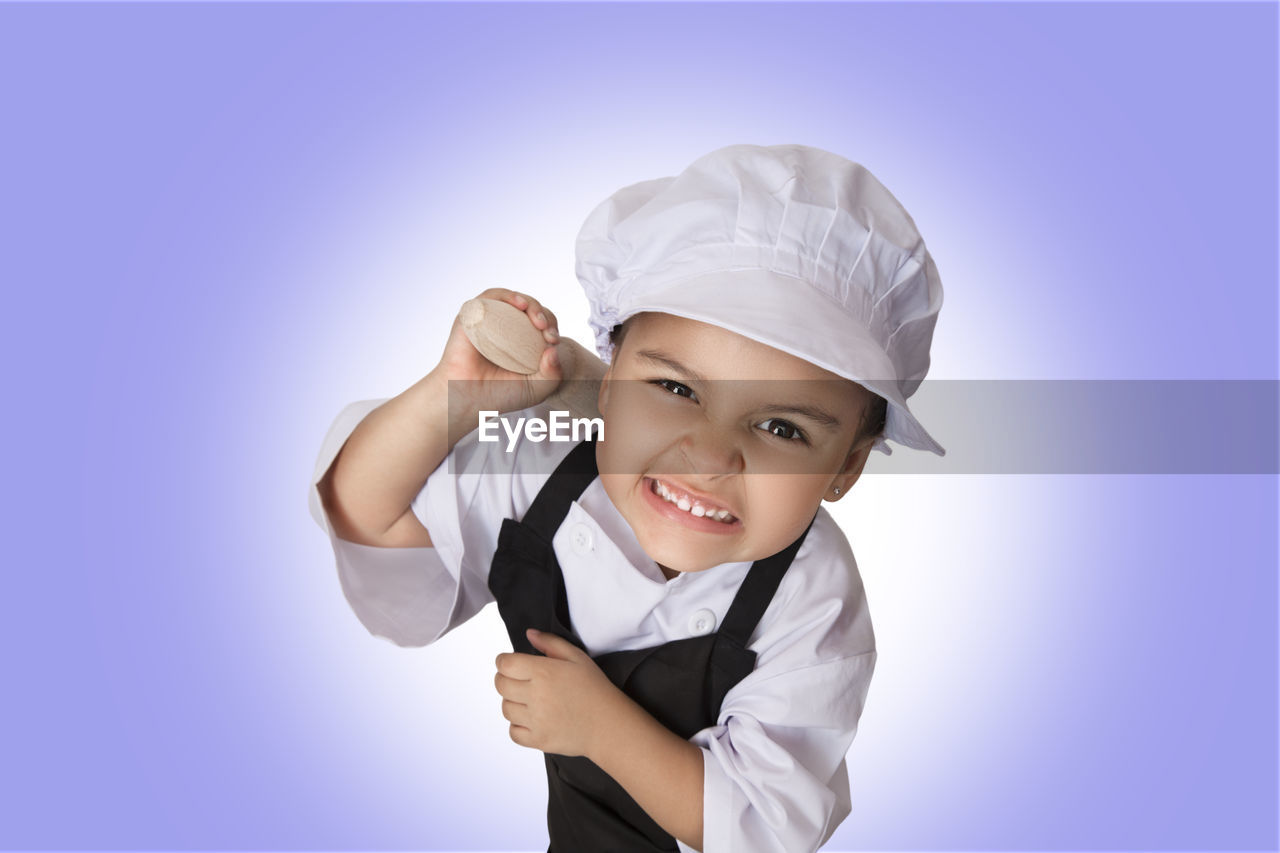 Portrait of girl in chef uniform holding rolling pin while standing against white background