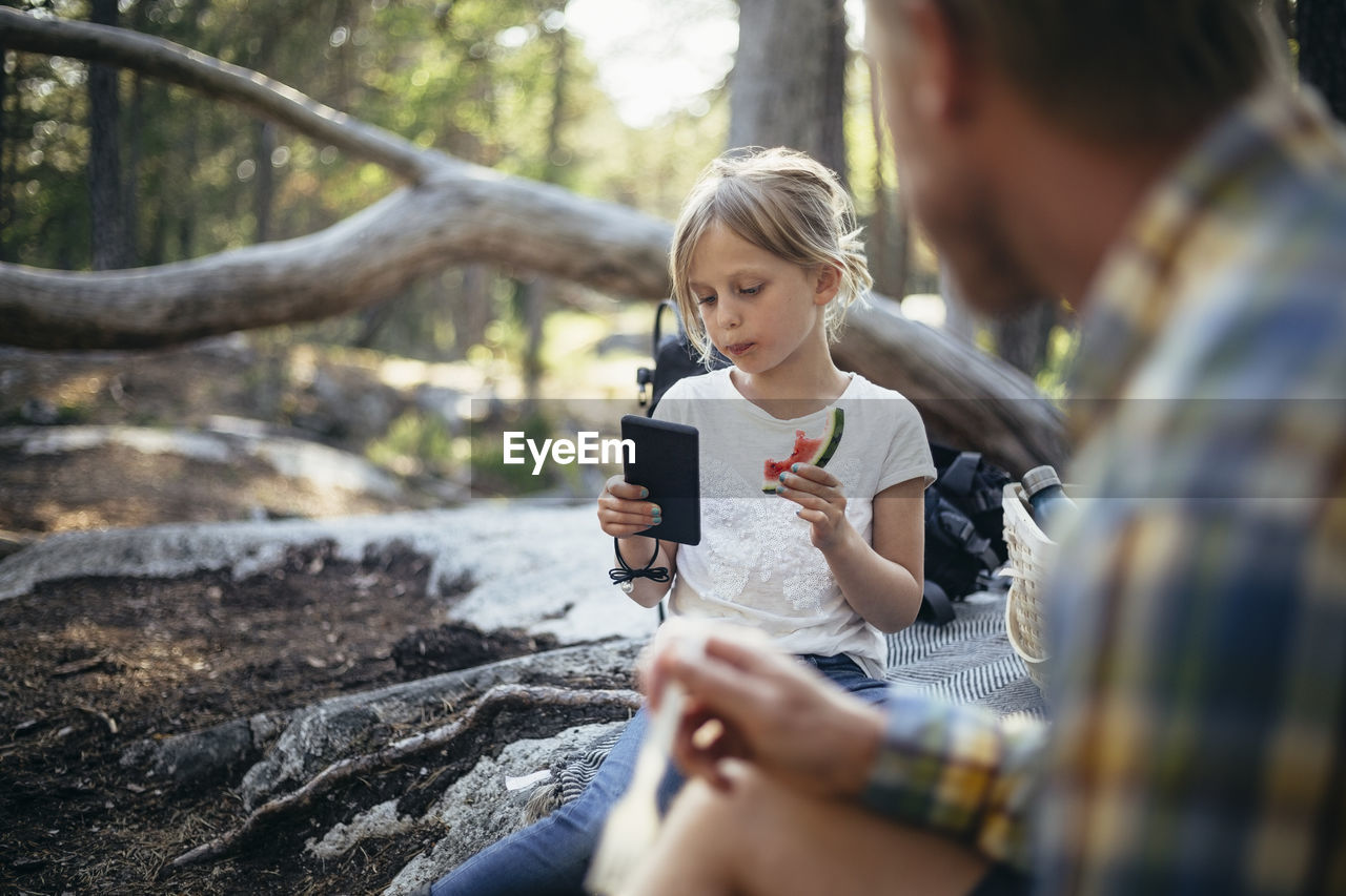 Daughter eating watermelon while using phone by father in forest