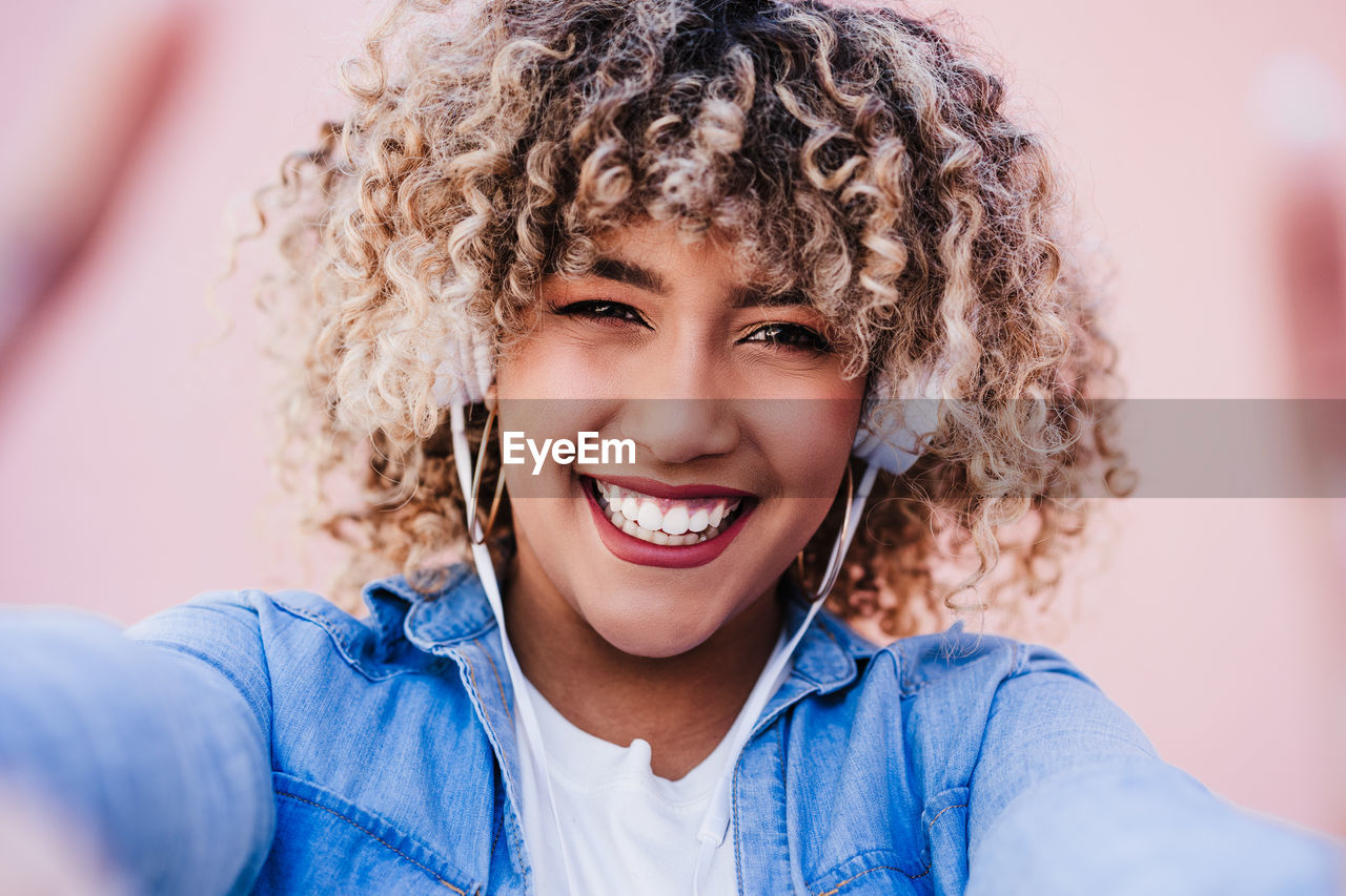 Portrait of smiling hispanic woman with afro hair in city using mobile phone and headset. lifestyle