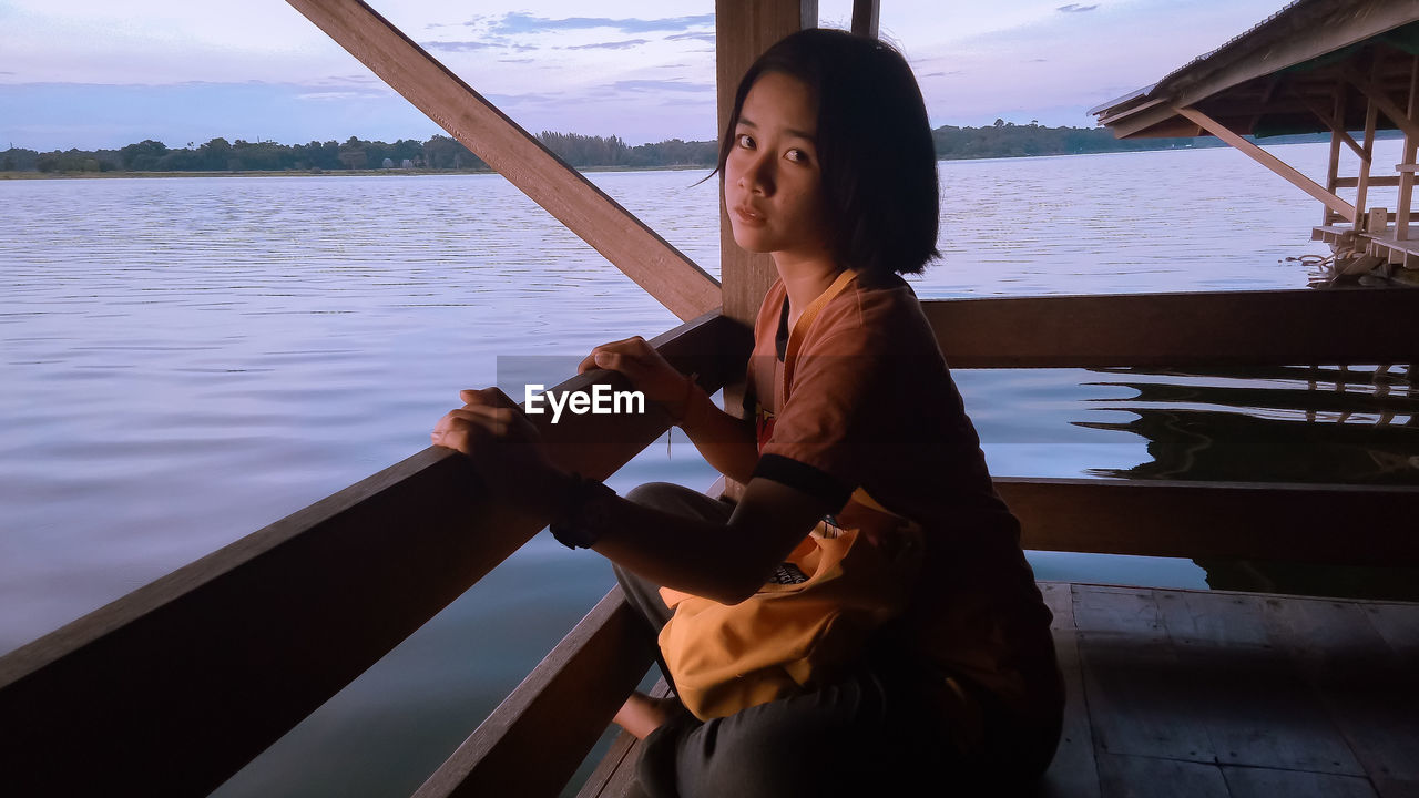 Portrait of young woman sitting on stilt house over lake against sky during sunset