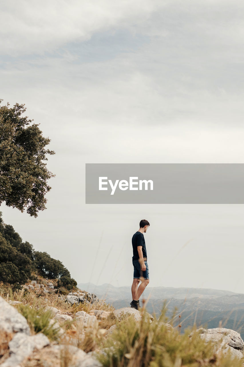 Full body side view of unrecognizable male hiker standing on rocky slope of mountain and observing landscape while travelling through highlands