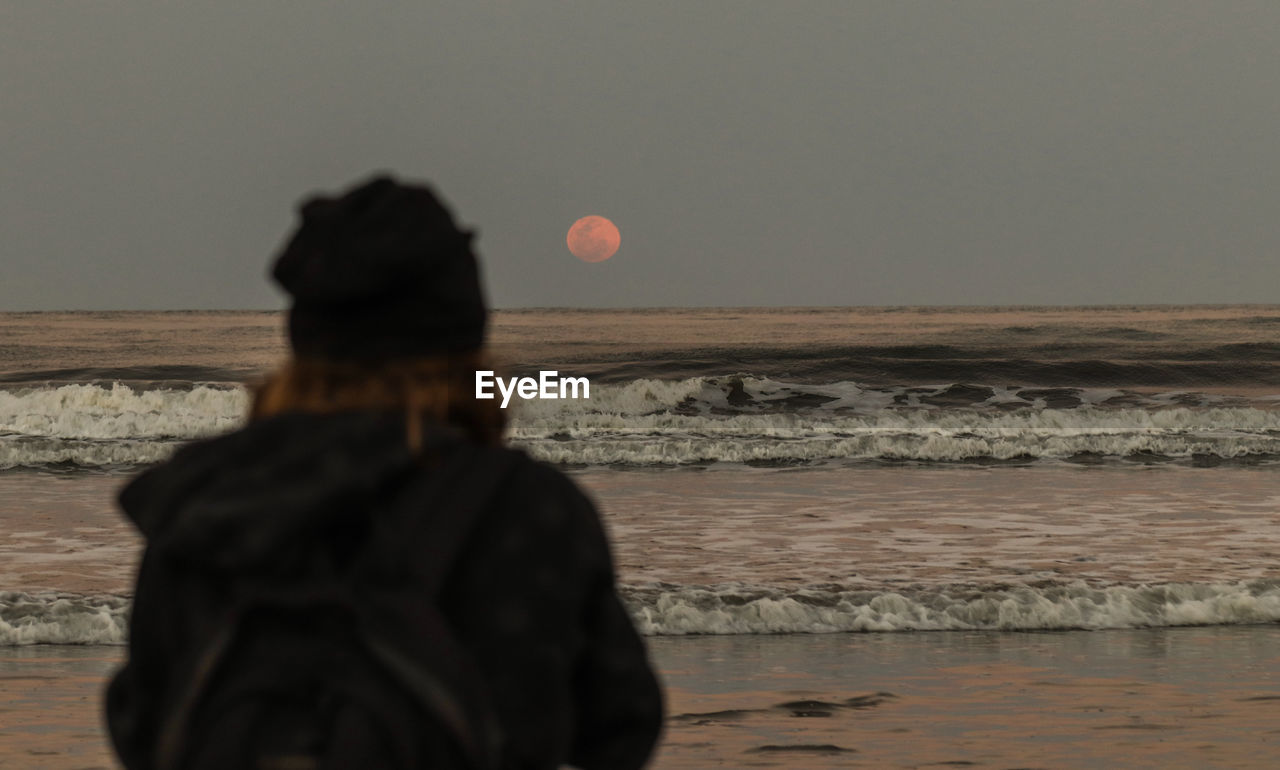 REAR VIEW OF WOMAN STANDING AT BEACH AGAINST SKY