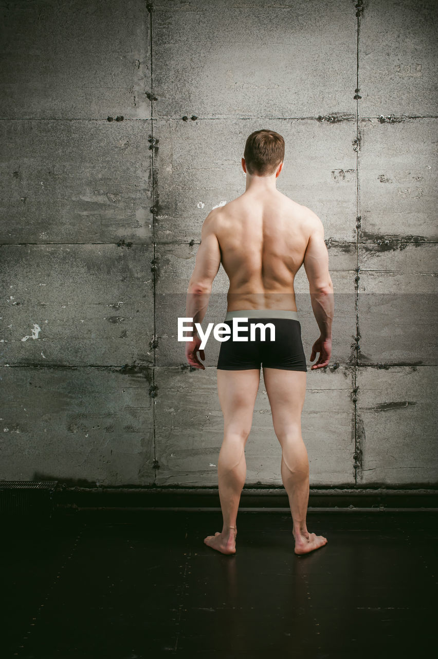 Rear view of shirtless muscular man standing by wall