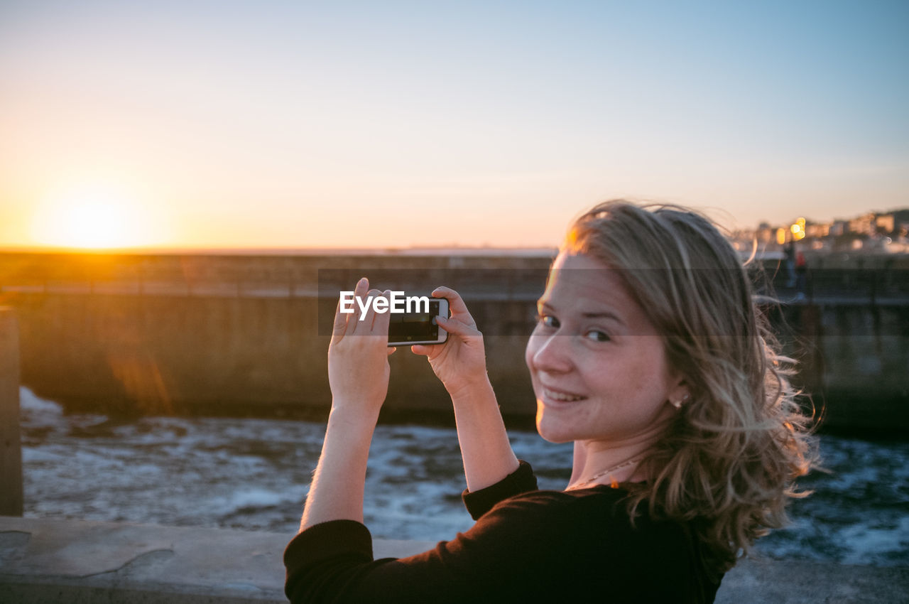 Portrait of smiling young woman photographing river during sunset