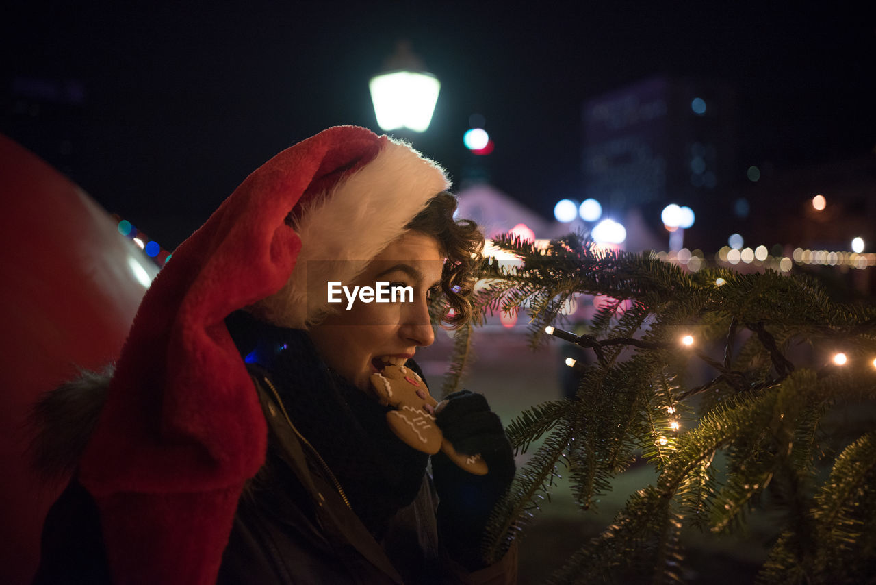 Young woman eating cookie by illuminated christmas tree at night