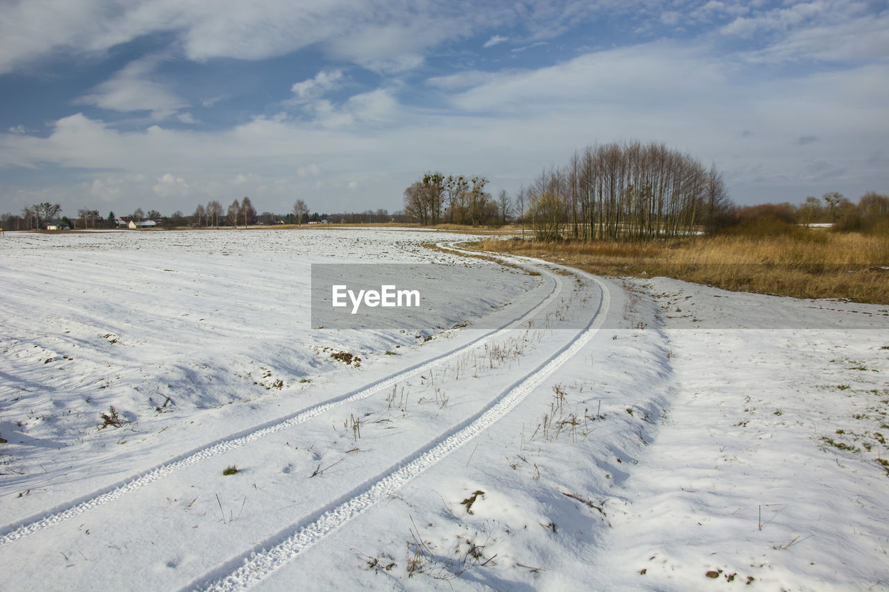 Traces of wheels on snowy dirt road, trees and clouds on blue sky