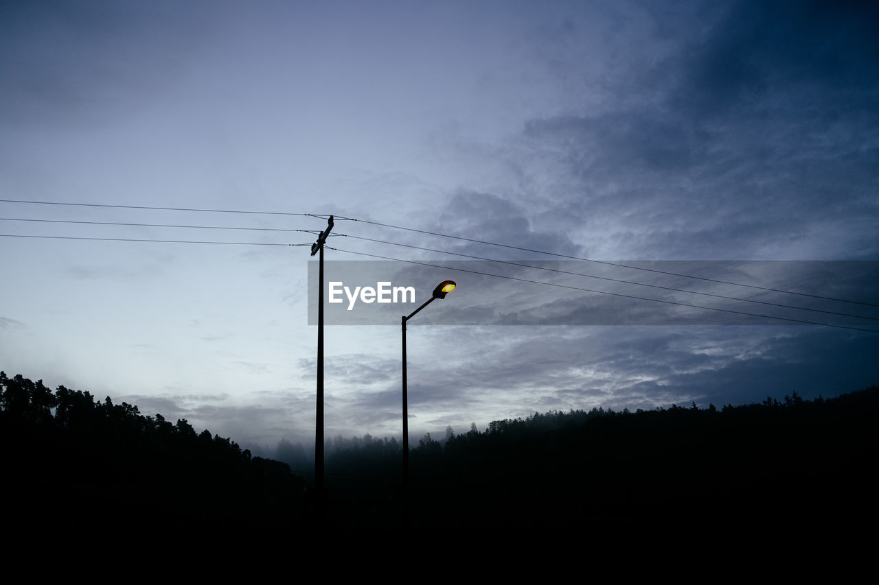 Yellow street light and telephone wire in front of morning sky and dark forest