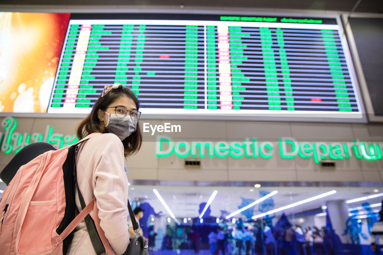 Woman tourist wearing mask standing with pink backpack and looking time board at airport