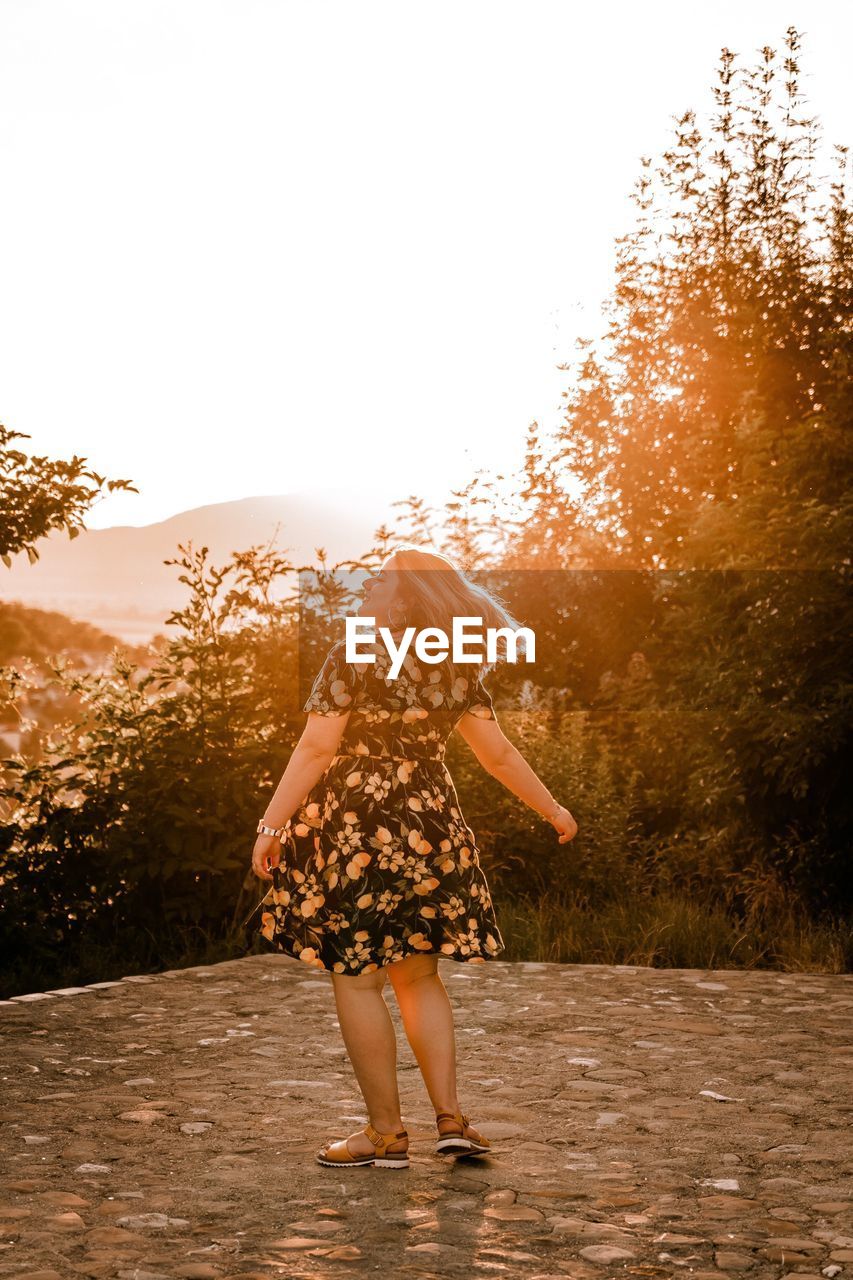 REAR VIEW OF WOMAN STANDING BY TREES AGAINST SKY