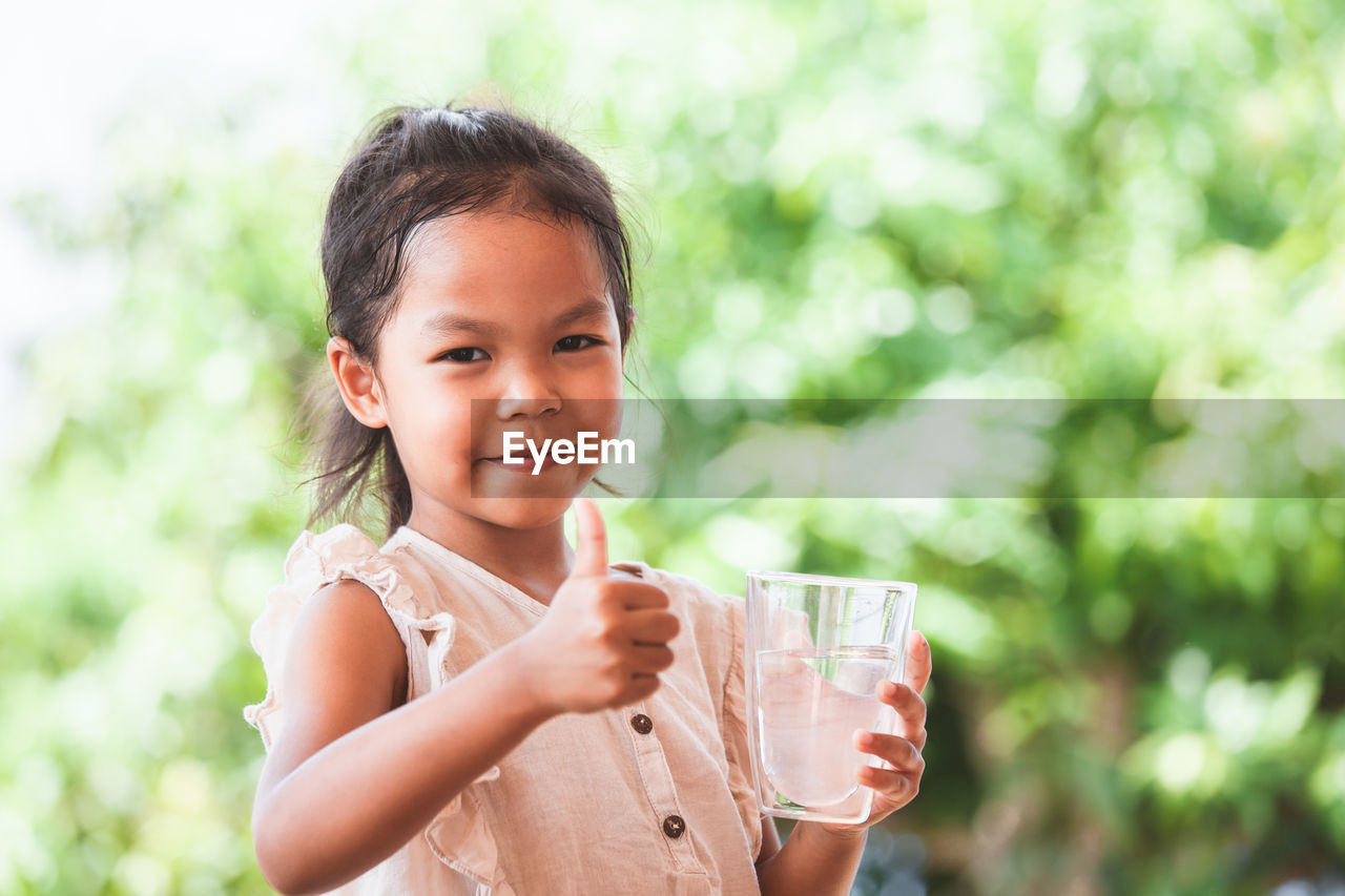 Portrait of girl gesturing thumbs up sign while holding water glass outdoors