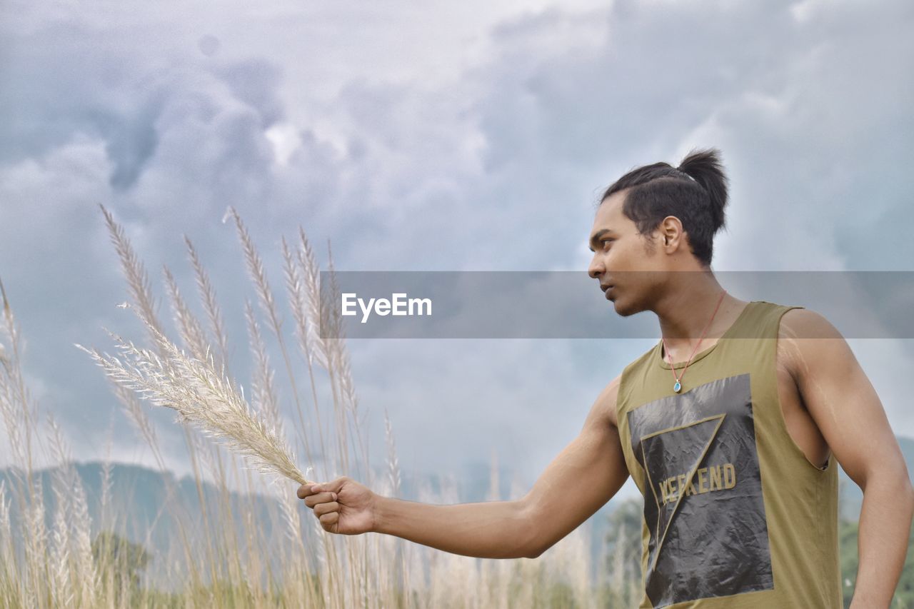 Young man holding plant while standing at farm against cloudy sky