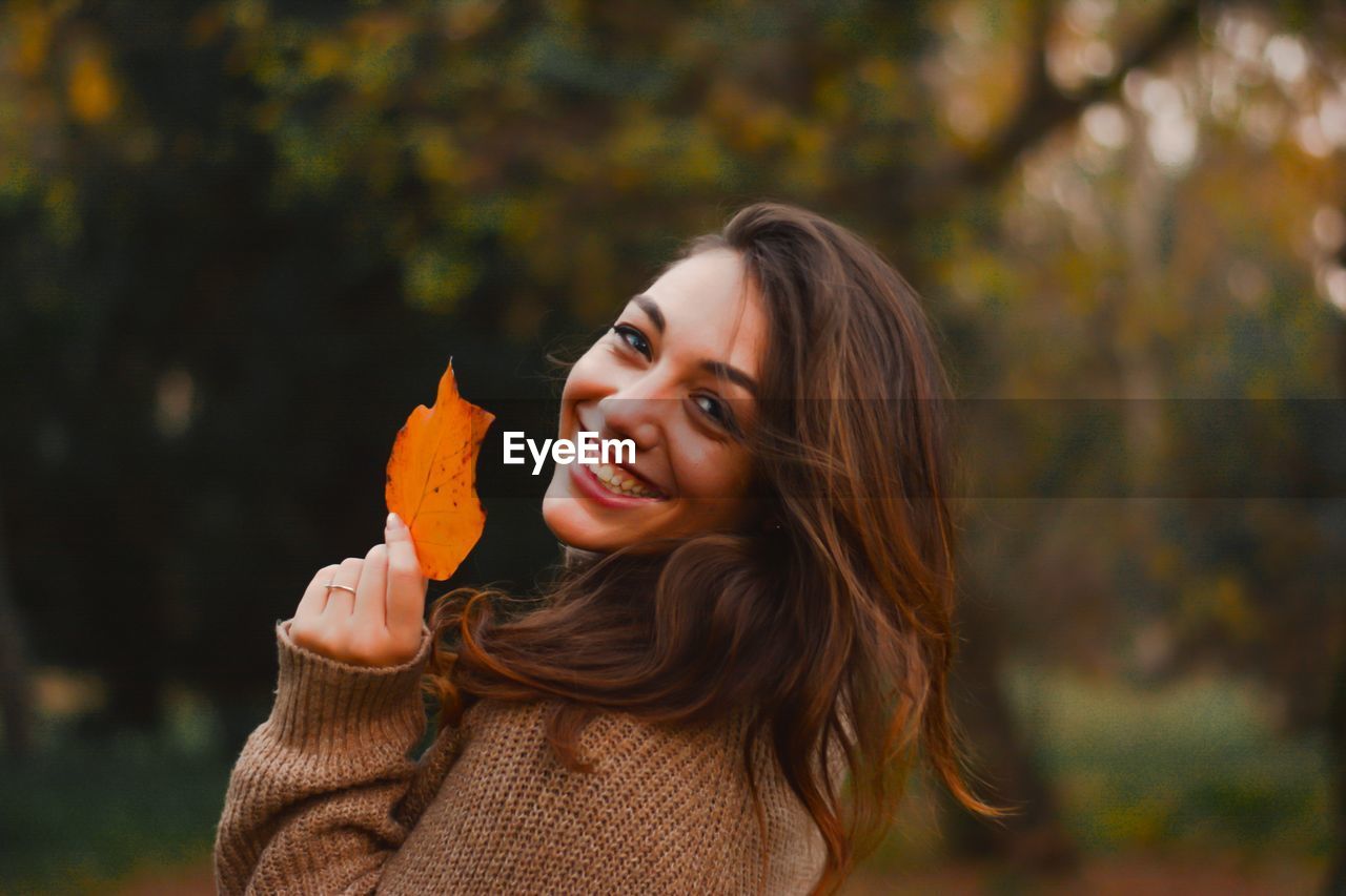 Portrait of smiling young woman holding autumn leaf during autumn at park