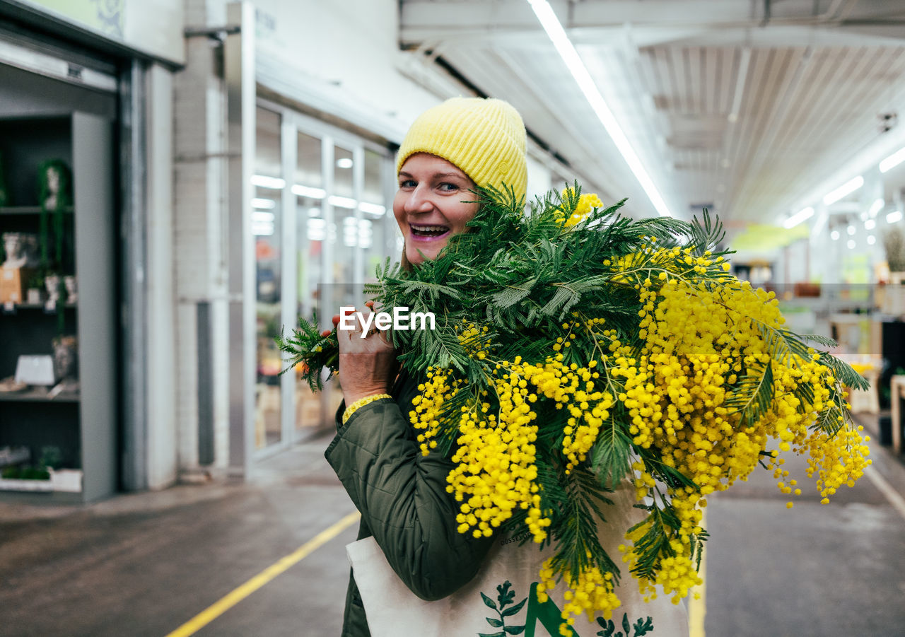 Positive woman in yellow cap and large bouquet of mimosa flowers at flower market