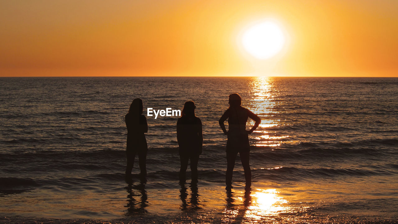 SILHOUETTE PEOPLE STANDING ON BEACH AGAINST ORANGE SKY