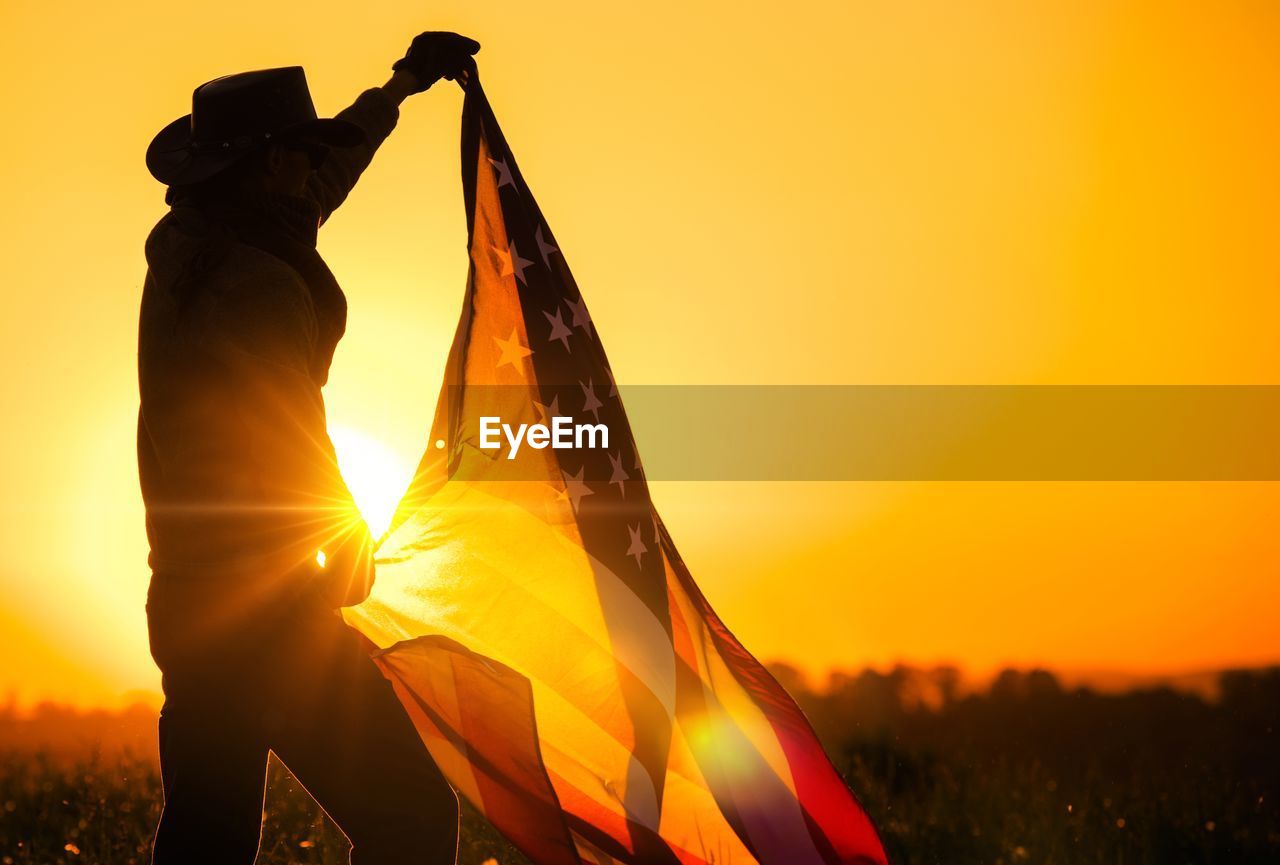 Silhouette man holding american flag on field against clear orange sky