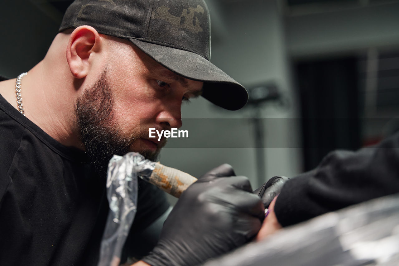 Concentrated male tattooist in gloves making tattoo on hand of client while using professional tattoo machine in modern tattoo studio