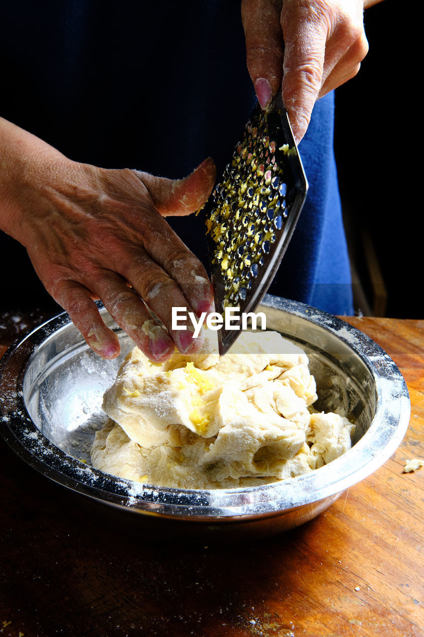 Unrecognizable female removing fresh lemon peel from metal grater over bowl with pastry dough in kitchen