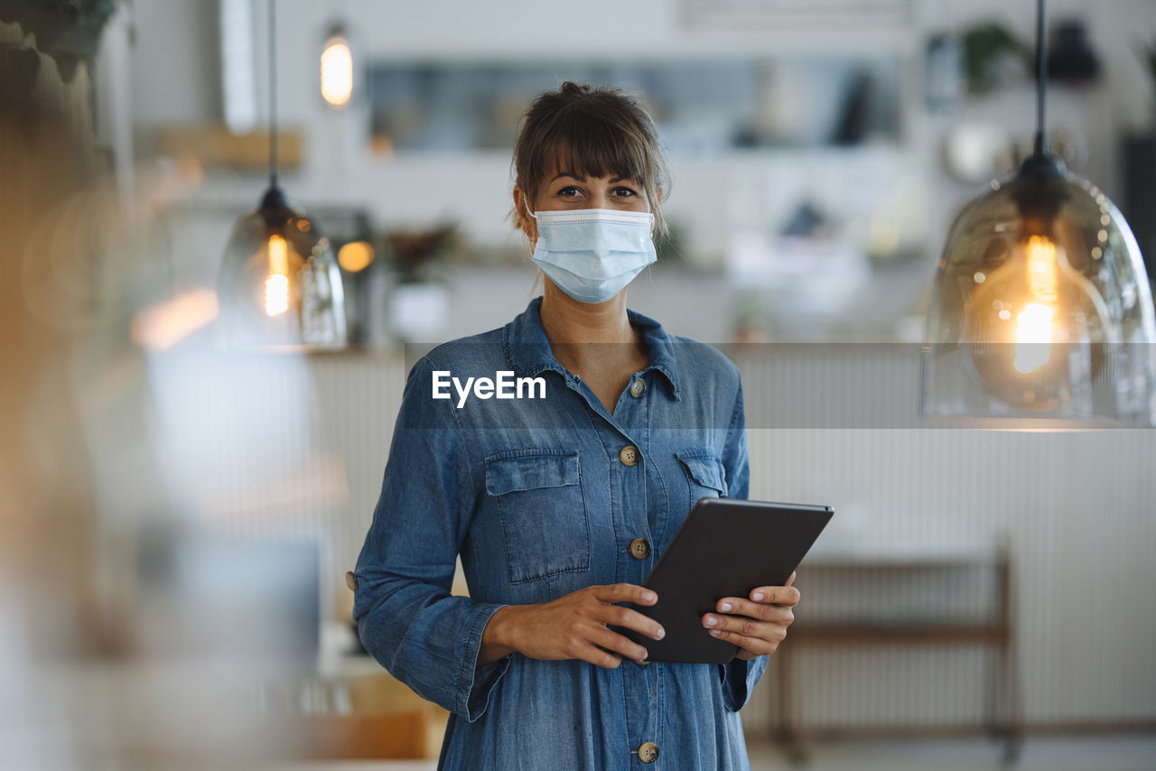 Female owner wearing protective face mask using digital tablet while standing in cafe