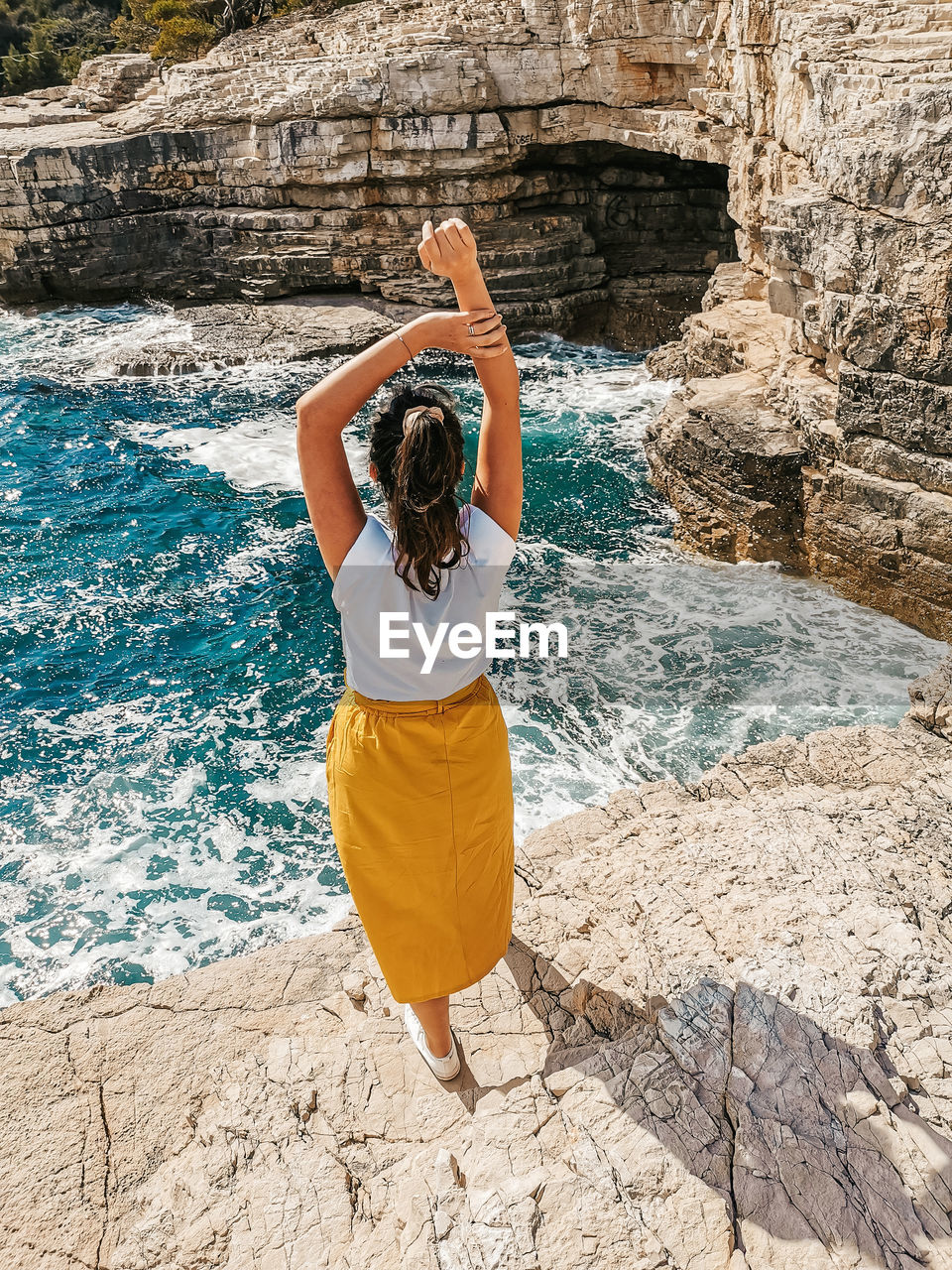 Young woman in yellow skirt standing on cliff above sea.