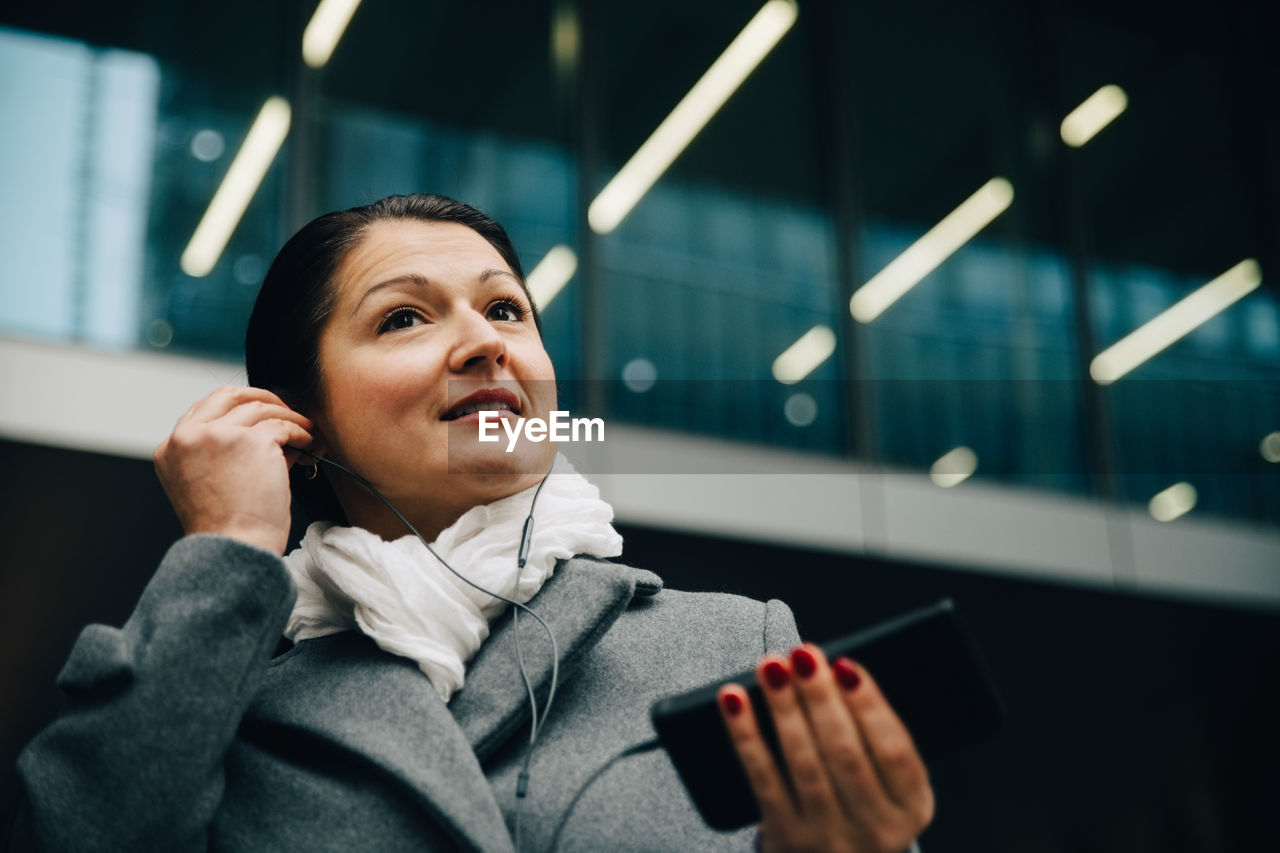 Low angle view of smiling businesswoman listening music while standing against building in city