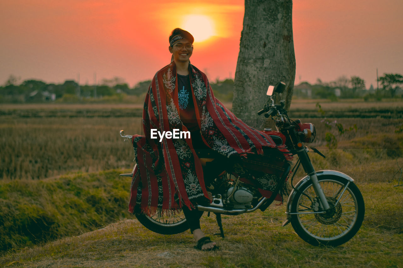 Portrait of smiling young man sitting on motorcycle at farm during sunset