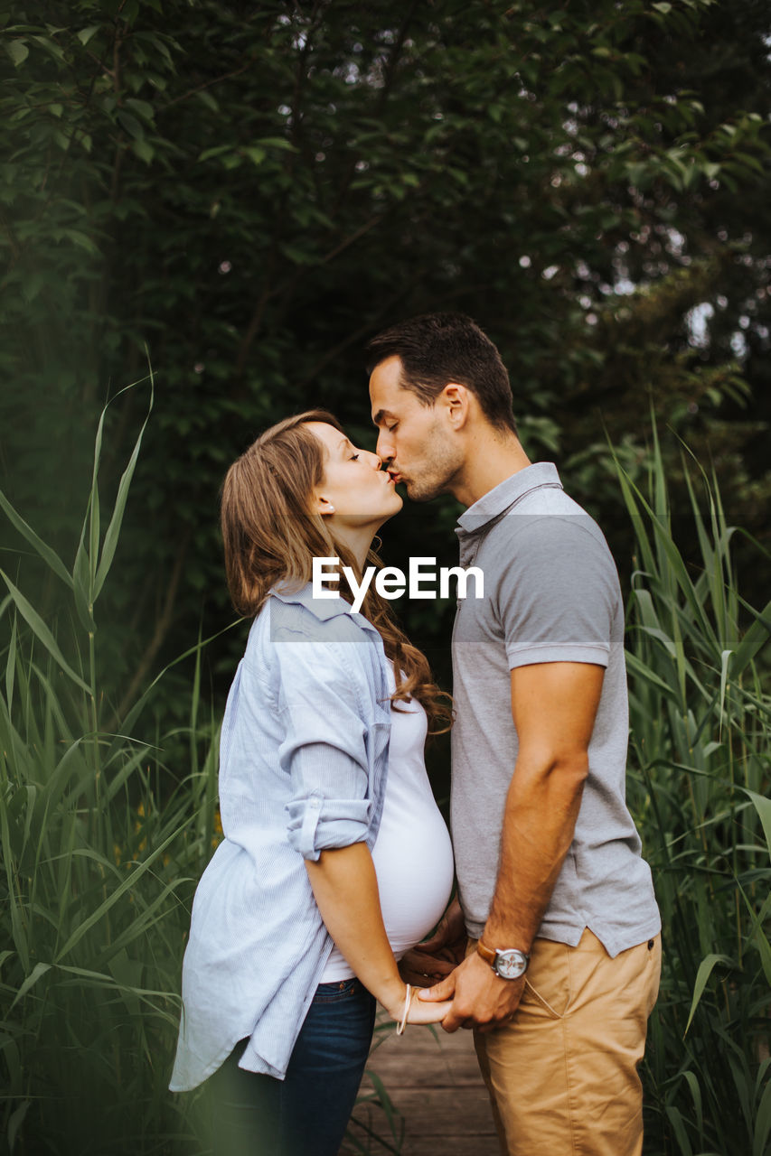 Side view of man kissing pregnant wife while standing amidst plants