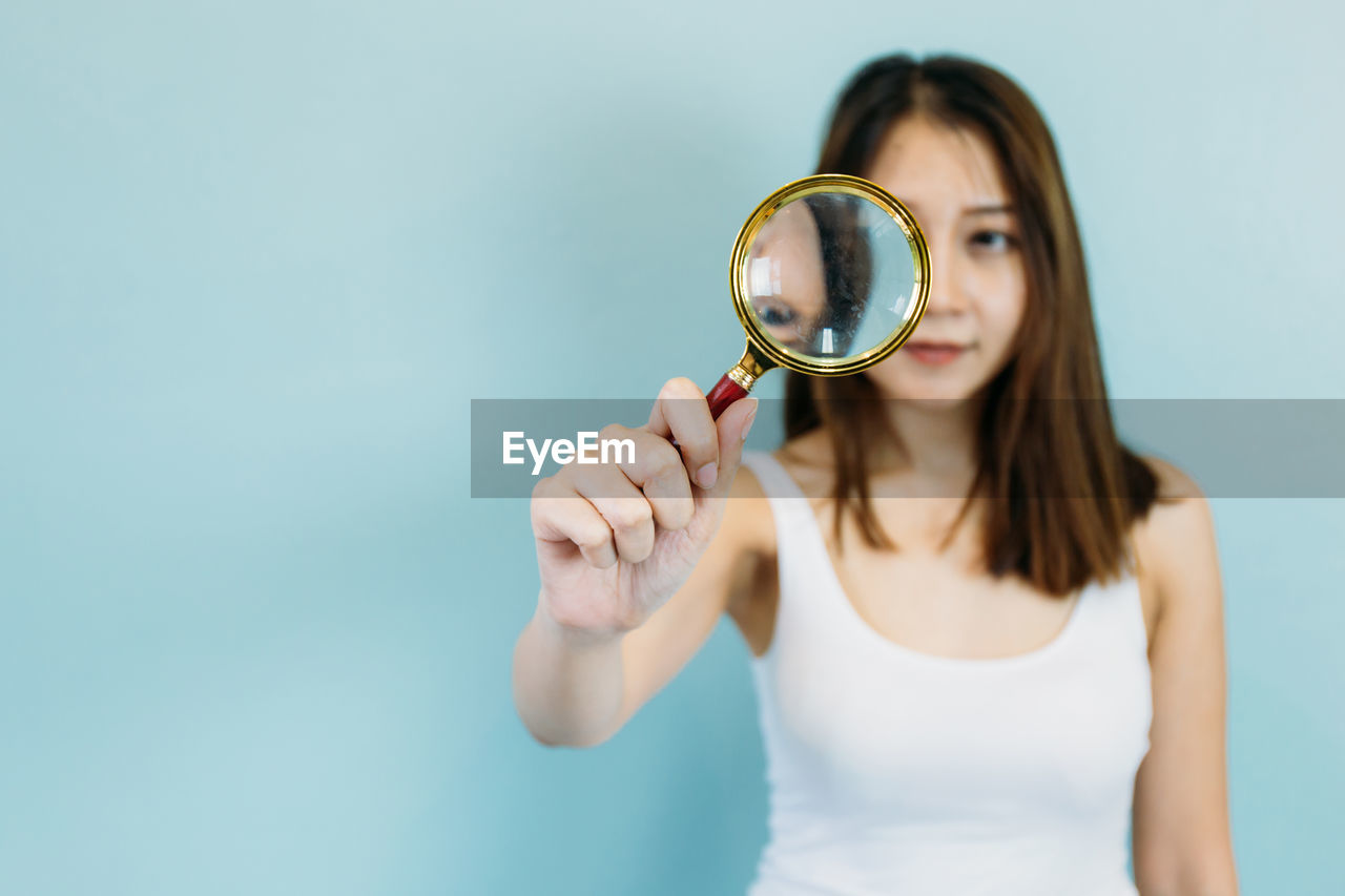 Close-up of young woman holding magnifying glass against blue background