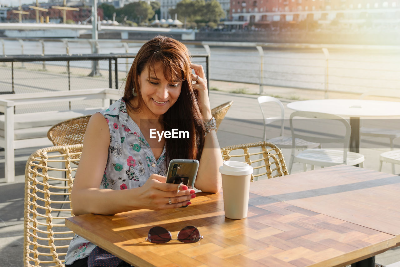 A hispanic woman looking her phone on coffee shop outside drinking coffee.