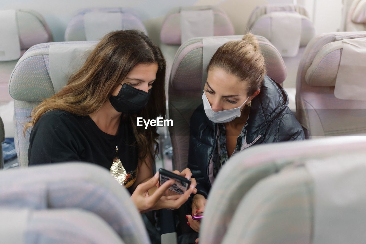 Woman with mask showing smartphone to friend in airplane