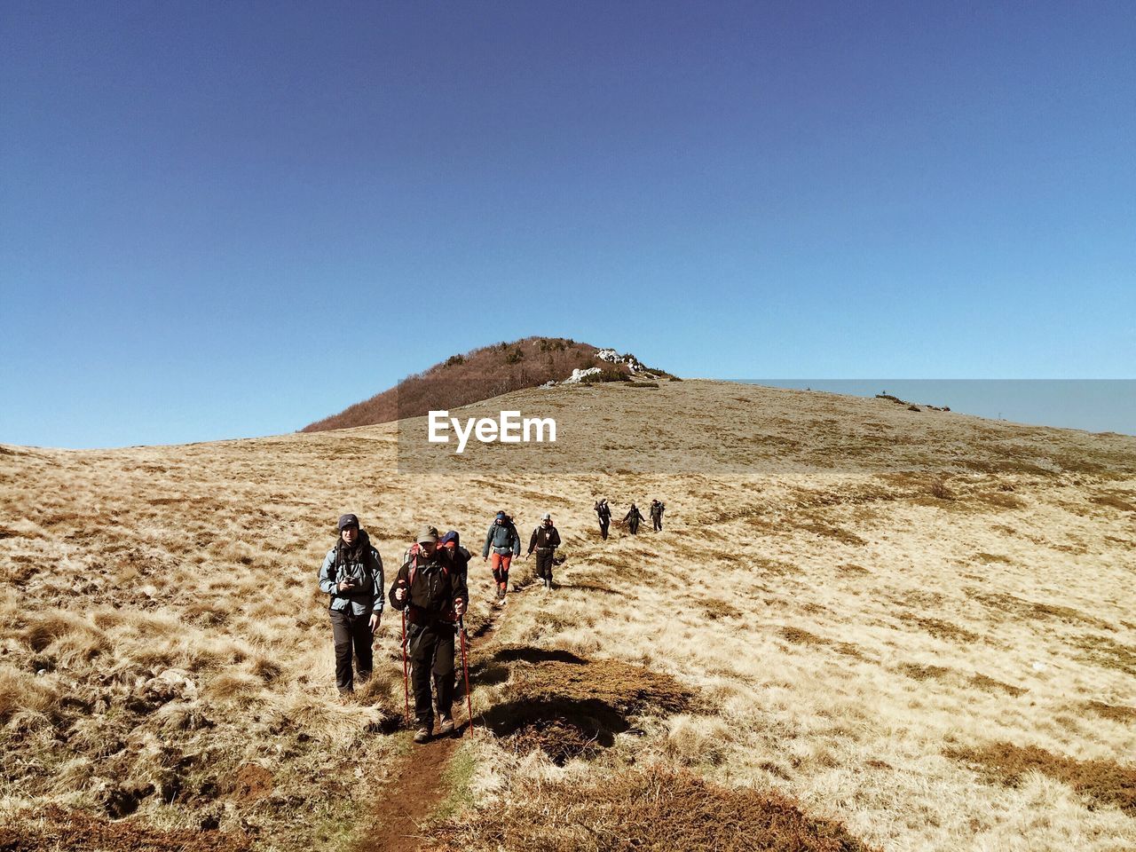 REAR VIEW OF PEOPLE WALKING ON ARID LANDSCAPE AGAINST CLEAR BLUE SKY