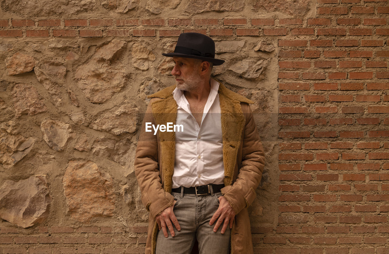 Portrait of adult man in hat and coat against yellow wall on street