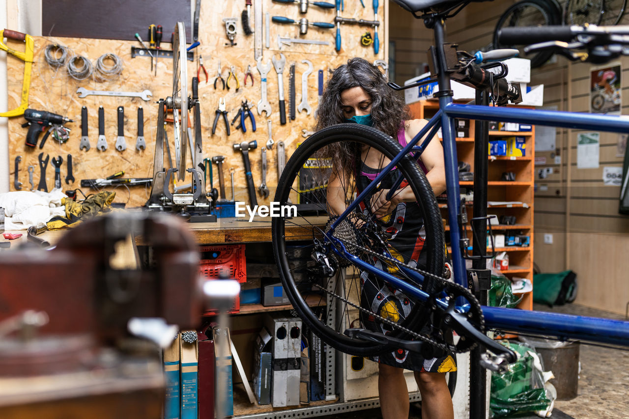 Side view of happy woman in workwear and protective face mask repairing wheel of bike during maintenance service in workshop