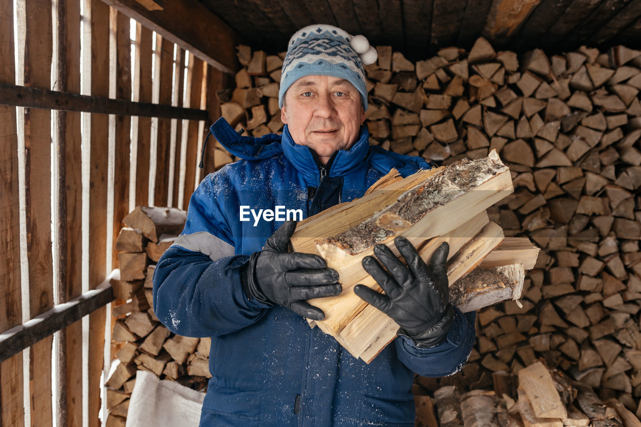 Senior farmer in a warm hat and jacket holds firewood against the background 