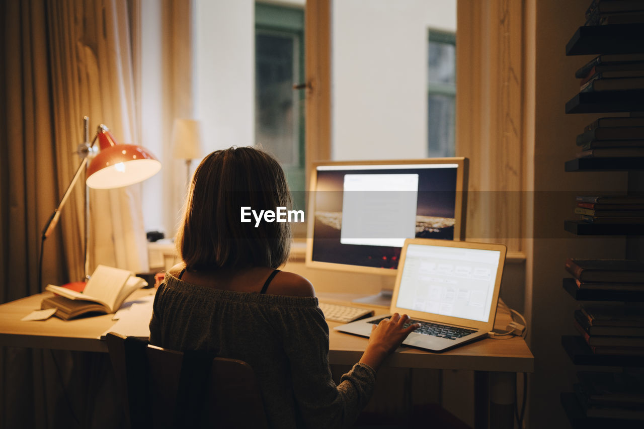 Rear view of girl using laptop and computer while sitting at illuminated desk